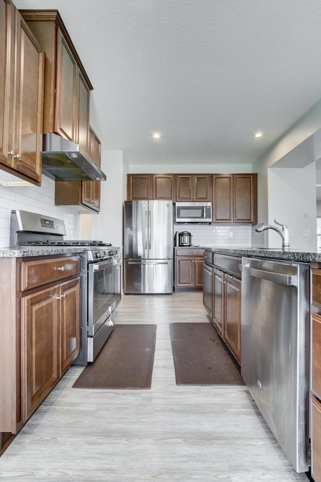 kitchen with stainless steel appliances, wall chimney range hood, a sink, light wood-style floors, and tasteful backsplash