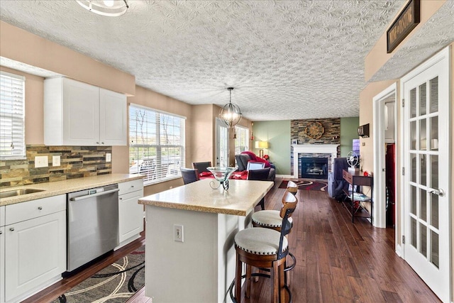 kitchen featuring white cabinetry, stainless steel dishwasher, a stone fireplace, a kitchen island, and dark wood-style floors