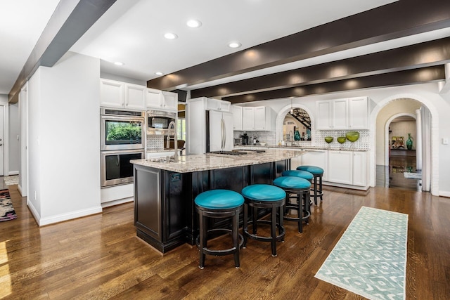 kitchen featuring decorative backsplash, appliances with stainless steel finishes, arched walkways, light stone counters, and white cabinets