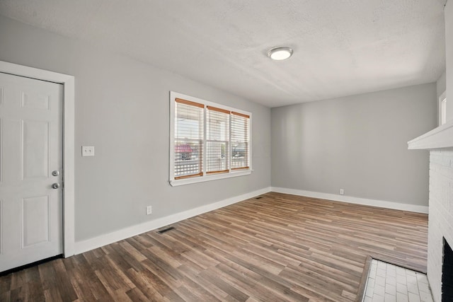 unfurnished living room with a textured ceiling, visible vents, baseboards, wood finished floors, and a brick fireplace