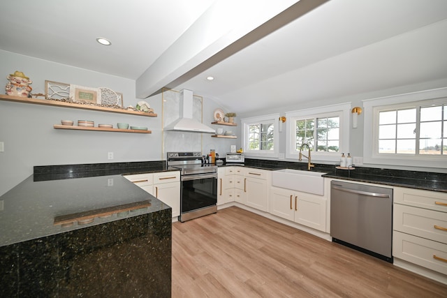 kitchen with dark stone countertops, open shelves, appliances with stainless steel finishes, a sink, and wall chimney exhaust hood