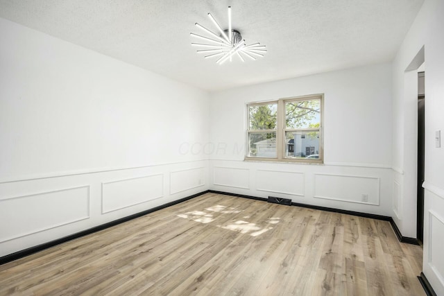 empty room featuring a decorative wall, wainscoting, light wood finished floors, a notable chandelier, and a textured ceiling