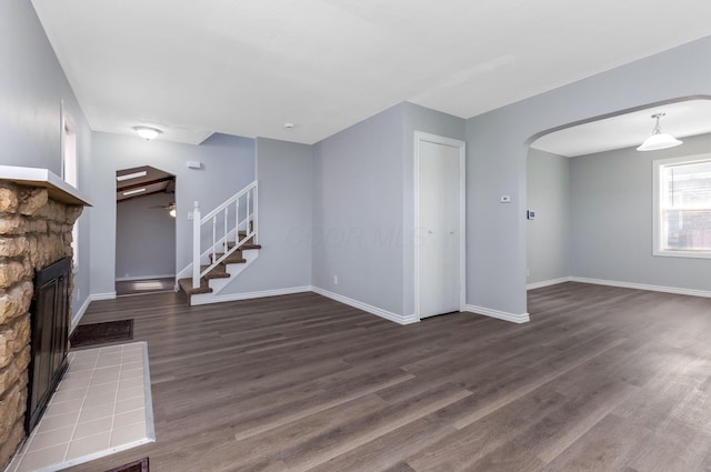 unfurnished living room featuring a ceiling fan, a stone fireplace, arched walkways, baseboards, and dark wood-type flooring