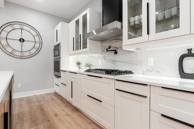 kitchen with wall chimney range hood, decorative backsplash, light wood-type flooring, white cabinets, and stainless steel appliances