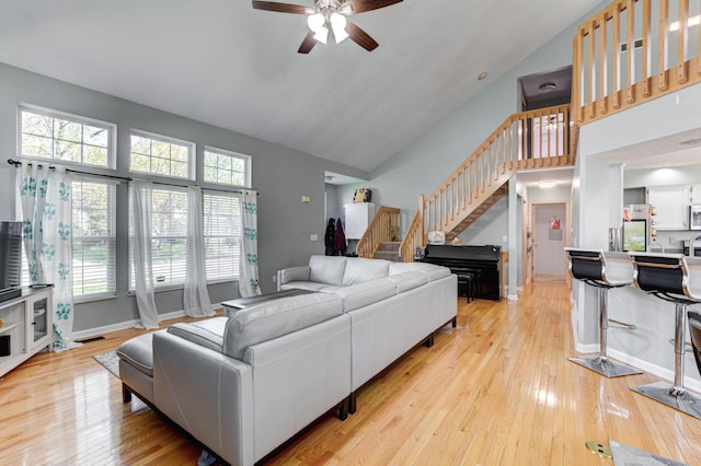 living room featuring stairway, a ceiling fan, light wood-style floors, baseboards, and high vaulted ceiling