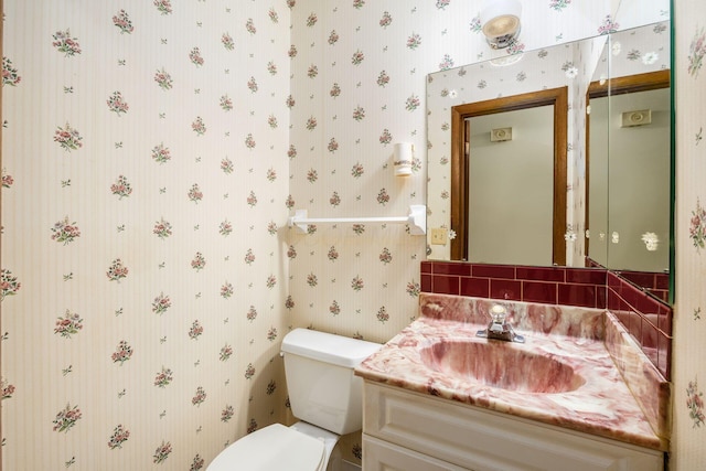half bathroom with toilet, vanity, and wallpapered walls