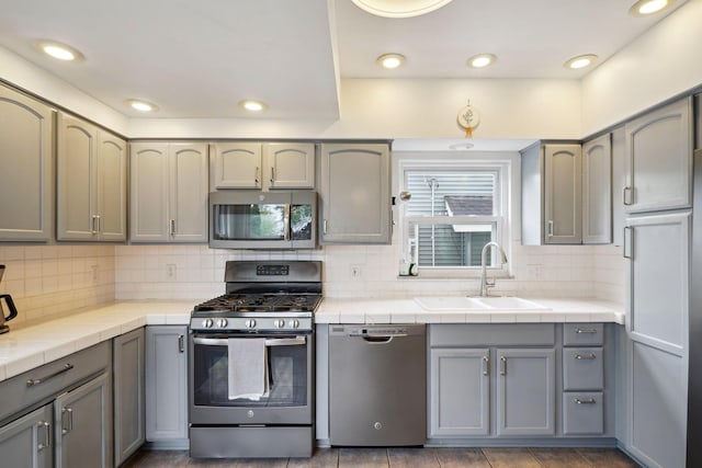 kitchen with appliances with stainless steel finishes, a sink, tile counters, gray cabinetry, and recessed lighting