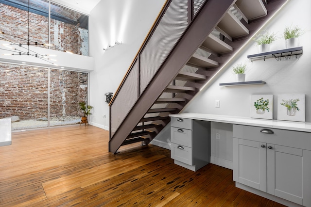 staircase featuring a high ceiling, brick wall, a chandelier, hardwood / wood-style floors, and baseboards