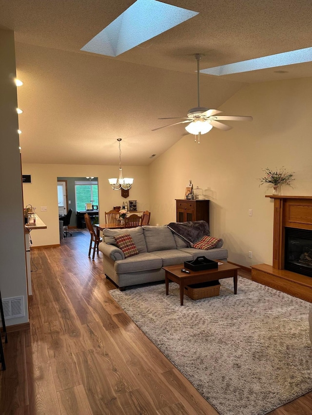 living area with visible vents, a fireplace with raised hearth, ceiling fan with notable chandelier, dark wood-style floors, and lofted ceiling with skylight