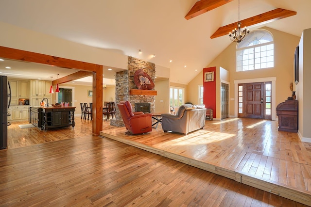 living room featuring a stone fireplace, beam ceiling, high vaulted ceiling, a notable chandelier, and light wood-style flooring