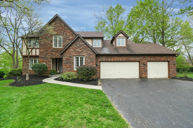 tudor-style house featuring a shingled roof, an attached garage, brick siding, a front yard, and driveway