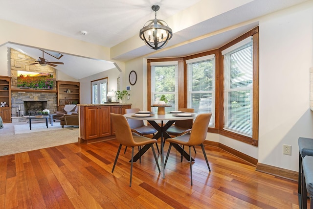 dining room featuring hardwood / wood-style flooring, ceiling fan with notable chandelier, a stone fireplace, baseboards, and lofted ceiling