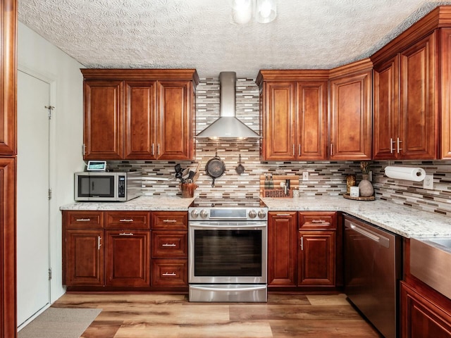 kitchen with wall chimney range hood, light stone counters, stainless steel appliances, light wood-type flooring, and backsplash