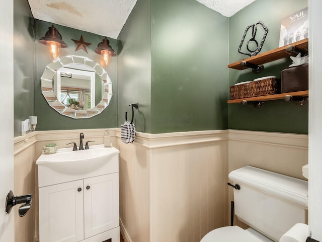 bathroom with a textured ceiling, vanity, toilet, and a wainscoted wall