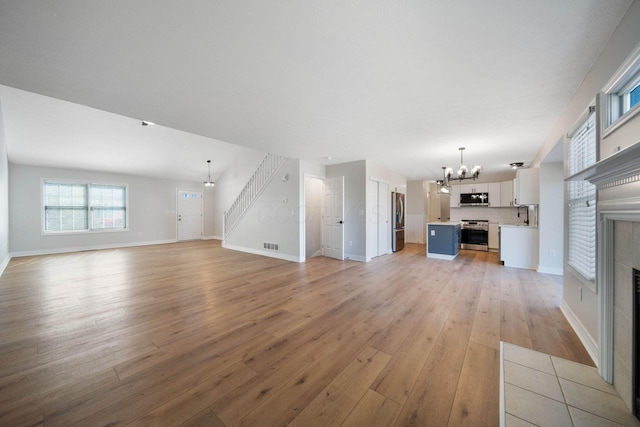 unfurnished living room with an inviting chandelier, visible vents, stairs, light wood-style floors, and a tiled fireplace