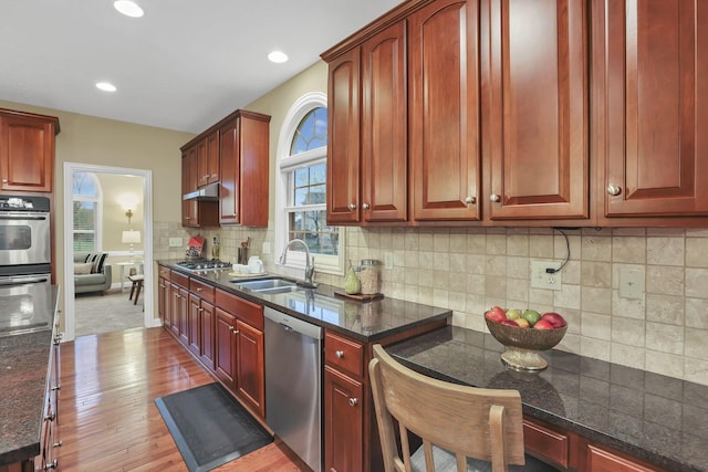 kitchen with wood finished floors, reddish brown cabinets, a sink, dark stone countertops, and stainless steel appliances