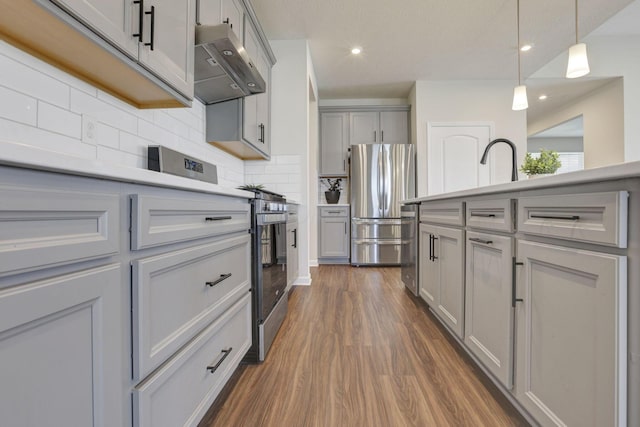 kitchen with wall chimney range hood, appliances with stainless steel finishes, gray cabinets, backsplash, and dark wood-style floors