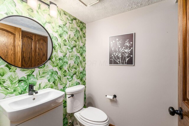 half bathroom with a textured ceiling, vanity, baseboards, toilet, and wallpapered walls