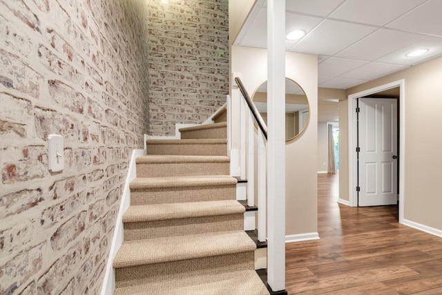 stairway with baseboards, brick wall, a paneled ceiling, and wood finished floors