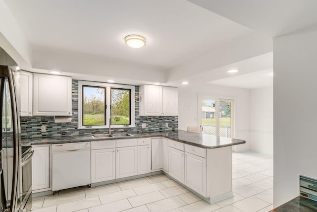 kitchen with white dishwasher, dark countertops, a sink, a peninsula, and tasteful backsplash
