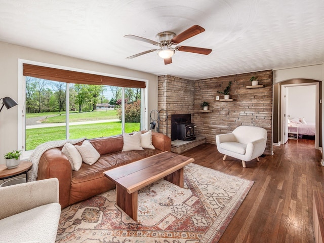 living area with a wood stove, ceiling fan, wood finished floors, and arched walkways