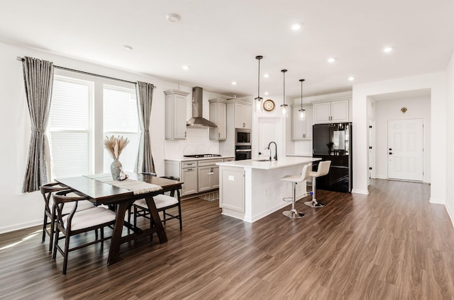 kitchen featuring a breakfast bar, a center island with sink, wall chimney exhaust hood, a sink, and stainless steel appliances