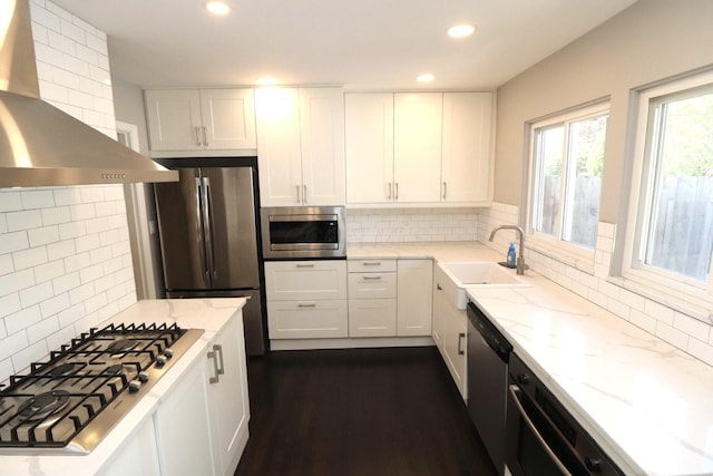 kitchen with wall chimney range hood, light stone countertops, a sink, stainless steel appliances, and backsplash
