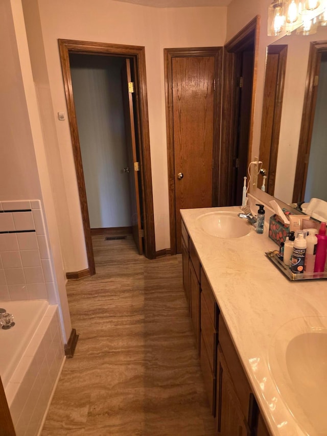 full bath featuring double vanity, wood finished floors, a sink, tiled bath, and baseboards