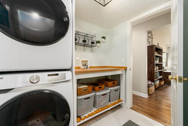 clothes washing area with laundry area, a textured ceiling, wood finished floors, stacked washing maching and dryer, and baseboards
