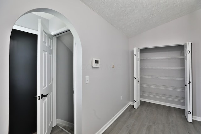 hallway featuring arched walkways, a textured ceiling, baseboards, wood finished floors, and lofted ceiling