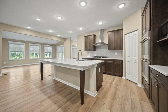 kitchen with a kitchen breakfast bar, stainless steel double oven, dark brown cabinets, a sink, and wall chimney exhaust hood
