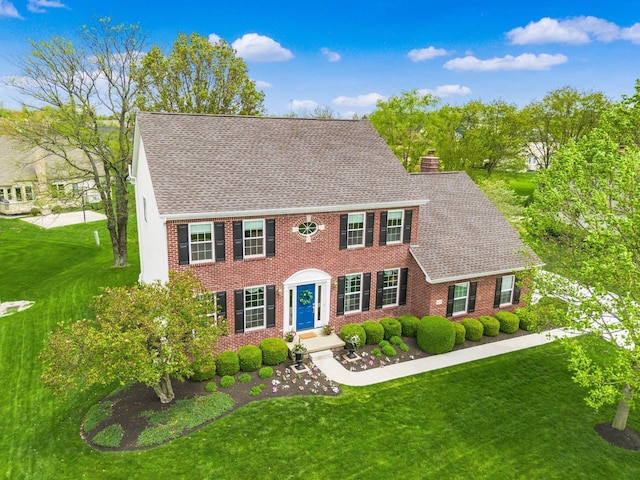colonial house featuring brick siding, a front lawn, roof with shingles, and a chimney