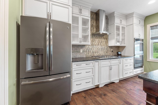 kitchen featuring wall chimney range hood, decorative backsplash, appliances with stainless steel finishes, dark wood-style floors, and glass insert cabinets