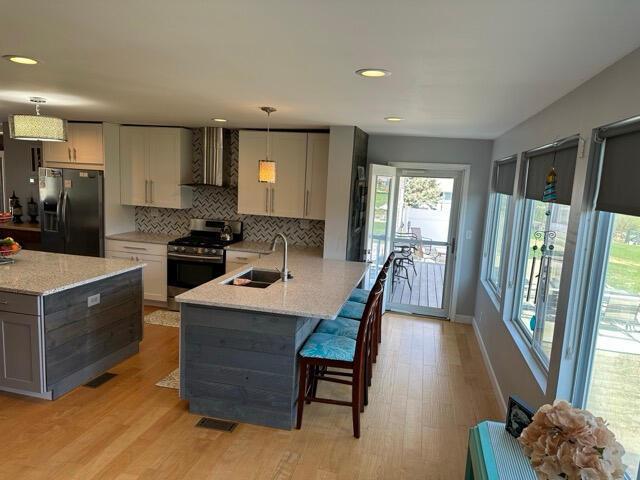 kitchen featuring wall chimney range hood, backsplash, stainless steel gas stove, a sink, and black refrigerator with ice dispenser
