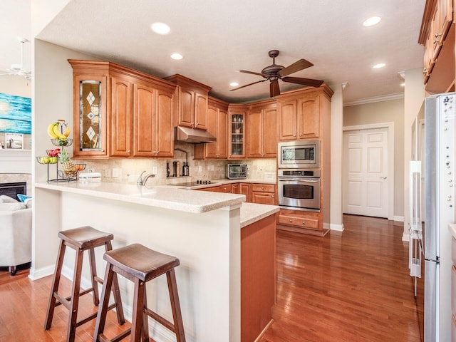 kitchen featuring a peninsula, stainless steel appliances, under cabinet range hood, a kitchen bar, and a ceiling fan