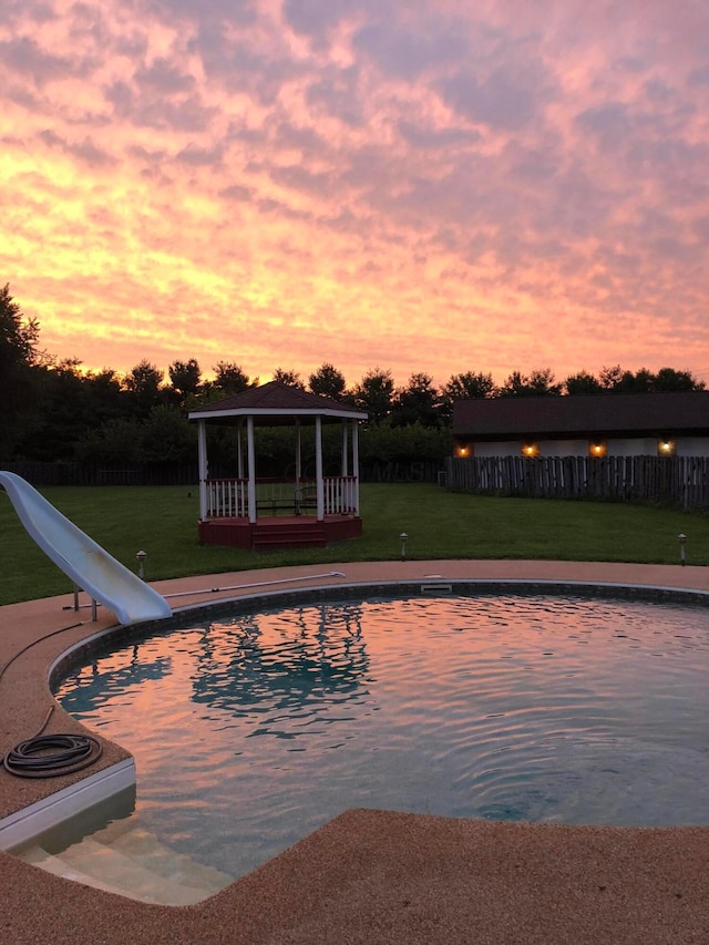 pool at dusk with a lawn, an outdoor pool, a gazebo, fence, and a water slide