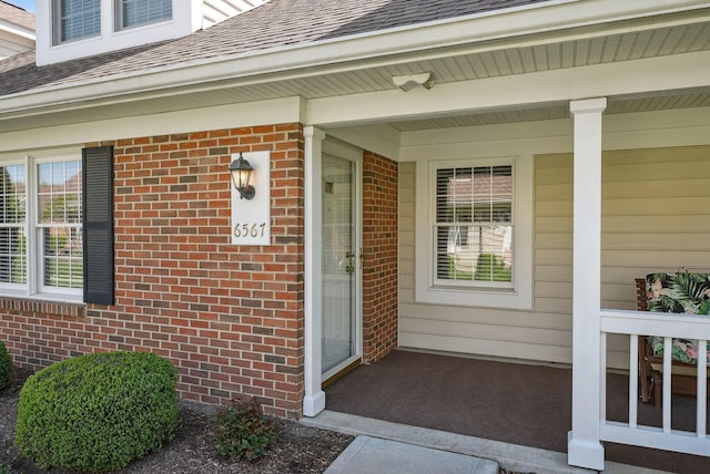 view of exterior entry featuring a shingled roof, a porch, and brick siding