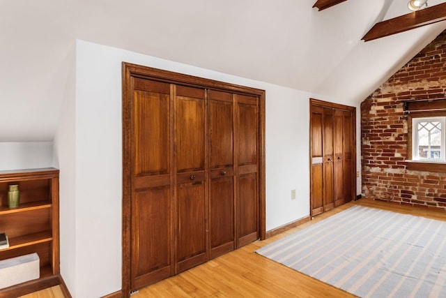 unfurnished bedroom featuring brick wall, baseboards, two closets, vaulted ceiling with beams, and light wood-style flooring