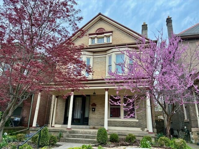 victorian home featuring covered porch, brick siding, and a chimney