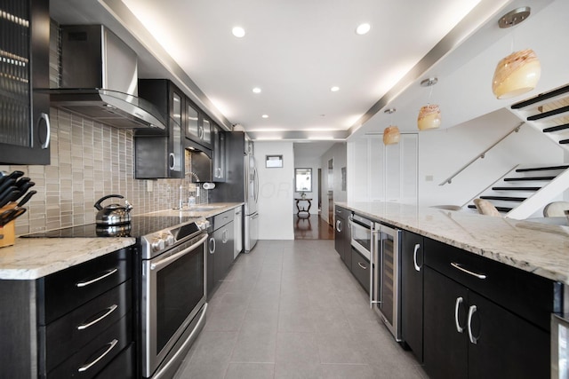 kitchen featuring wall chimney range hood, decorative backsplash, appliances with stainless steel finishes, glass insert cabinets, and dark cabinetry