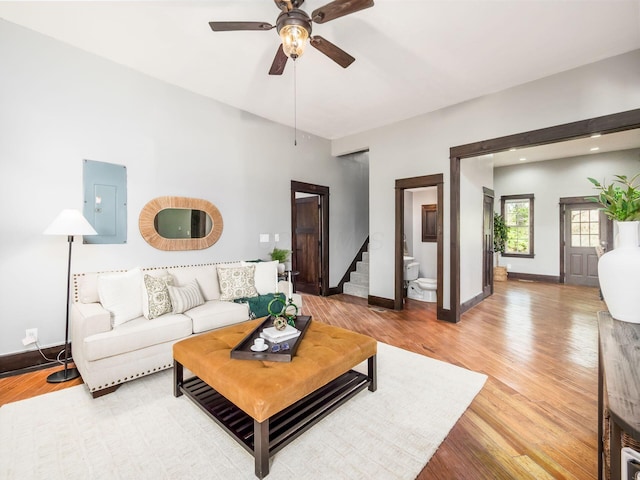 living room featuring stairs, a ceiling fan, baseboards, light wood-type flooring, and electric panel
