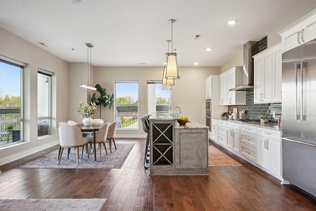 kitchen with built in appliances, wall chimney exhaust hood, a center island with sink, white cabinetry, and light stone counters