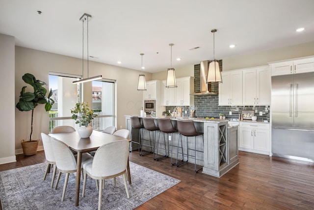 kitchen featuring built in appliances, wall chimney range hood, a kitchen island with sink, tasteful backsplash, and white cabinetry