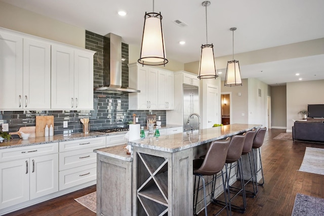 kitchen with wall chimney range hood, decorative backsplash, white cabinets, a kitchen island with sink, and a sink
