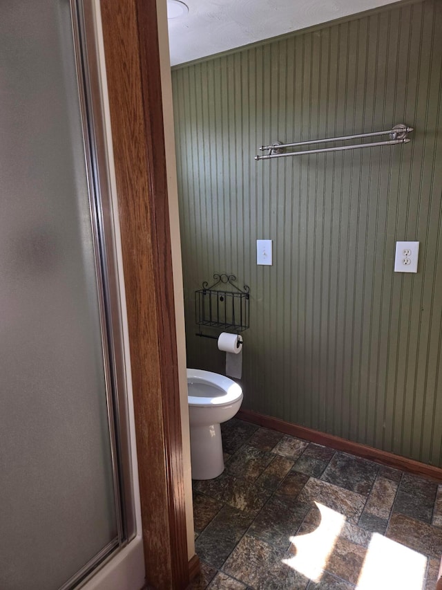 bathroom featuring wooden walls, baseboards, toilet, and stone finish flooring