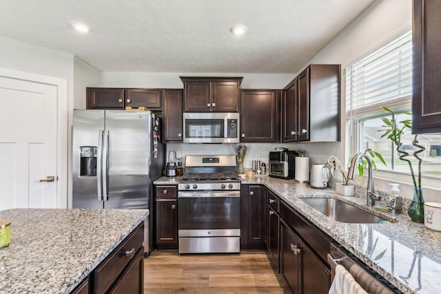 kitchen with dark brown cabinets, light stone countertops, appliances with stainless steel finishes, a sink, and light wood finished floors