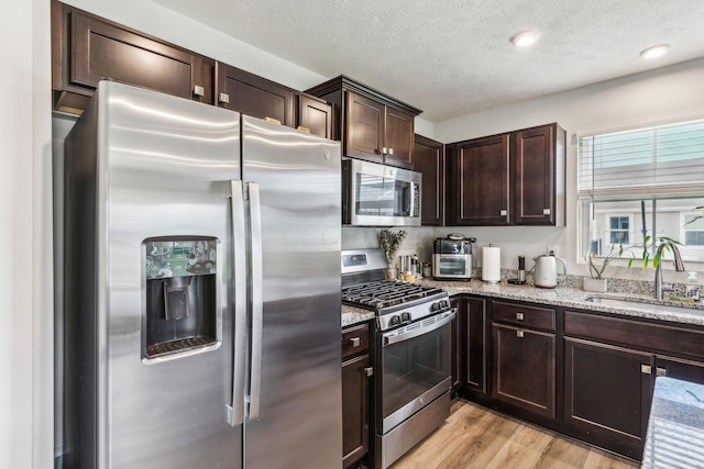 kitchen with appliances with stainless steel finishes, dark brown cabinets, a sink, light stone counters, and light wood-style floors
