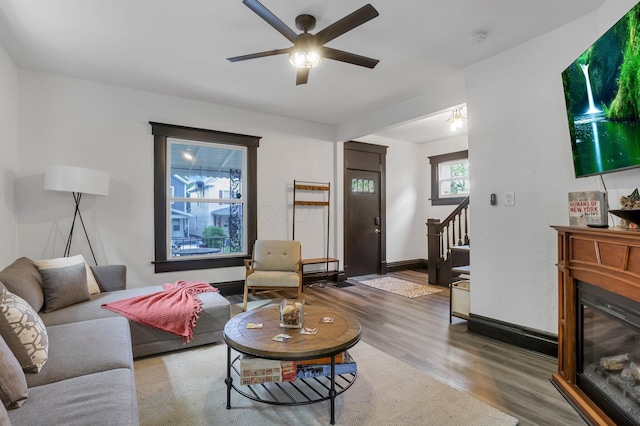 living area with ceiling fan, wood finished floors, baseboards, a glass covered fireplace, and stairs