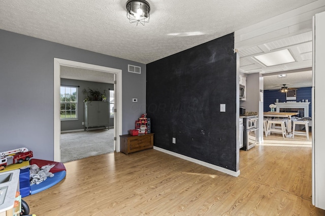 recreation room with visible vents, a brick fireplace, light wood-type flooring, a textured ceiling, and a ceiling fan