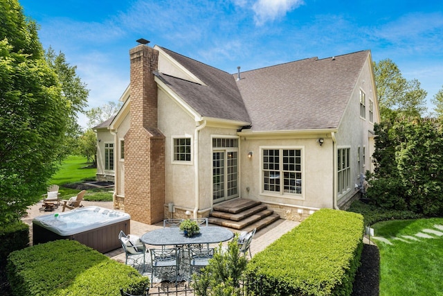 rear view of house with a chimney, roof with shingles, stucco siding, a hot tub, and a patio area